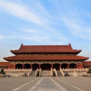 Un tradizionale palazzo cinese dal tetto di tegole rosse si erge alla fine di un ampio e vuoto cortile in pietra, sotto un cielo blu.
