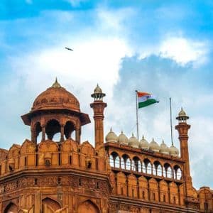 La bandera india ondea sobre una fortaleza ornamentada de arenisca roja con múltiples cúpulas y torres contra un cielo azul nublado.