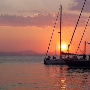 Diverse barche a vela ormeggiate in un porto, sagomate contro un tramonto arancione che si riflette sull'acqua.