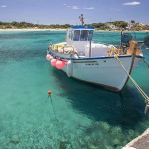 Una barca da pesca bianca con boe rosa ormeggiata in acqua cristallina turchese vicino a un molo in una giornata di sole.