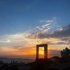 Un viaggio di gruppo WeRoad osserva il sole tramontare attraverso un grande arco di pietra in riva al mare sotto un cielo colorato.