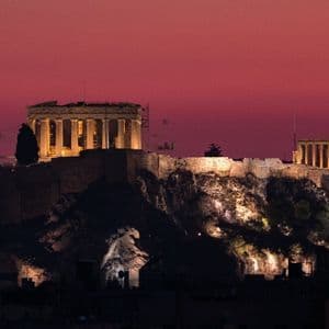 Due antichi templi con colonne sono illuminati su una collina rocciosa contro un cielo rosso intenso al tramonto.