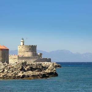Un mulino a vento in pietra con tetto arancione e un faro fortificato si ergono su una costa rocciosa accanto al mare blu, con montagne in lontananza.