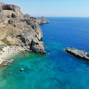 Una vista aerea di una piccola spiaggia incastonata in una cala rocciosa, con rovine sulla scogliera e barche nel mare limpido e turchese.