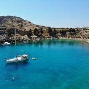 Una barca a vela bianca ancorata in una baia di acqua turchese, con una costa rocciosa e una piccola spiaggia sullo sfondo.
