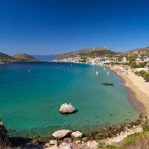 A high-angle view of a sandy beach and coastal town bordering a bay of clear turquoise water, with hills in the background.