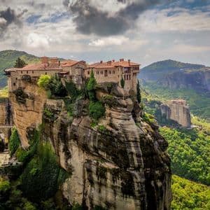 A stone monastery with a red-tiled roof sits atop a tall, sheer rock pillar, overlooking a vast, green, mountainous valley.
