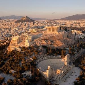 Aerial view of the Acropolis and a Roman amphitheater in Athens, illuminated by golden hour light, overlooking the cityscape.