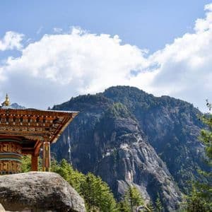 Una ruota di preghiera colorata in un padiglione di legno su una roccia, che si affaccia su una montagna boscosa con un monastero sulla parete della scogliera.