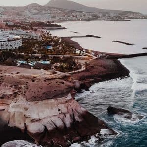 Una vista aerea di una città costiera al tramonto, con edifici lungo una costa rocciosa e spiagge di sabbia scura accanto all'oceano.