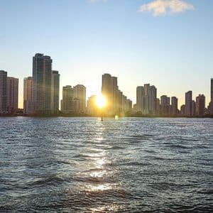 Eine Skyline hoher Gebäude, vom Wasser aus bei Sonnenuntergang gesehen, mit der Spiegelung der Sonne auf den Wellen.
