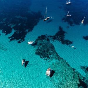 Una vista aerea di diverse barche a vela e catamarani che galleggiano in acqua trasparente turchese sopra un fondale sabbioso e roccioso.