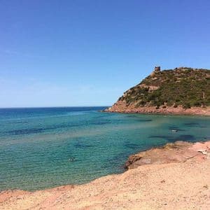 Una torre di pietra su una collina verde si affaccia su una baia calma con acqua turchese, una barca a vela e persone che nuotano da una spiaggia sabbiosa.