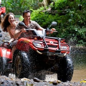 A WeRoad group trip smiles while riding red ATVs through a shallow stream in a lush jungle, splashing water.