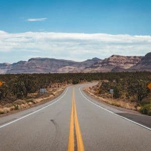 Una strada a due corsie serpeggiante che attraversa un paesaggio desertico con alberi di Joshua e montagne rocciose sotto un cielo parzialmente nuvoloso.