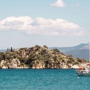 White boats float on turquoise water in front of a small, rocky island covered in greenery under a cloudy sky.