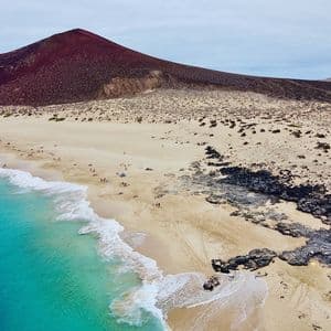 Eine Luftaufnahme eines Sandstrandes mit türkisfarbenem Wasser und vulkanischen Felsen, mit einem großen, roten Berg im Hintergrund.