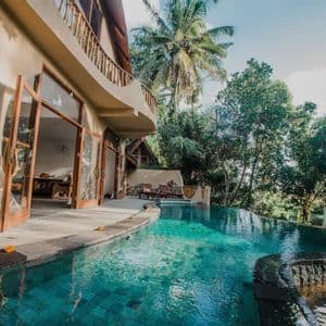 Una piscina a sfioro con acqua turchese accanto a una villa moderna con porte a vetro aperte, circondata da una rigogliosa foresta tropicale.