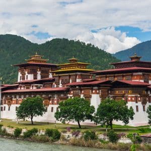 Un grande e tradizionale monastero bianco con tetti ornati di rosso e giallo si trova accanto a un fiume, alla base di verdi montagne boscose.