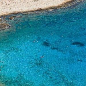 Vista aerea di una costa con spiaggia sabbiosa e persone che nuotano in acqua cristallina turchese.