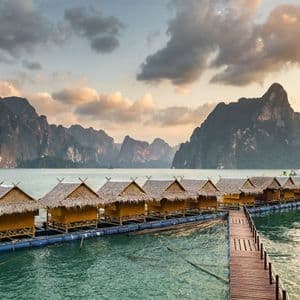 Eine Reihe schwimmender Bungalows mit Strohdächern auf einem ruhigen See, vor einer Kulisse dramatischer Berge unter einem bewölkten Himmel.