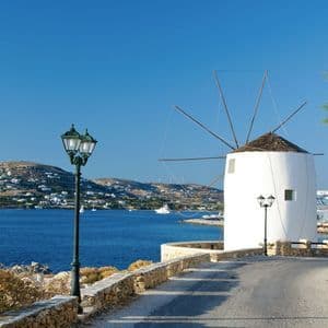 Un tradizionale mulino a vento bianco si erge su una strada costiera che domina una luminosa baia azzurra con colline e case in lontananza.