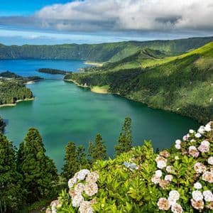 Una vista dall'alto di un grande lago vulcanico circondato da colline verdi, con ortensie bianche in primo piano sotto un cielo nuvoloso.