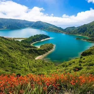 Una vista panoramica da una collina con fiori arancioni in primo piano, che si affaccia su un lago vulcanico di un blu brillante circondato da montagne verdi.