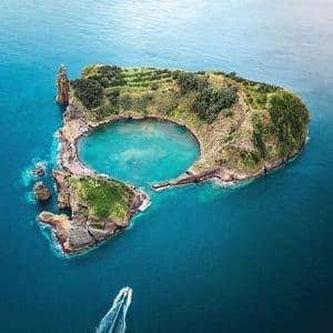 Vista aerea di un isolotto vulcanico verde con una laguna centrale turchese, mentre una motobarca passa nel mare blu profondo.