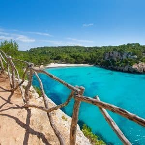 Une clôture en bois rustique longe un sentier côtier, surplombant une crique isolée aux eaux turquoises et une plage de sable blanc.