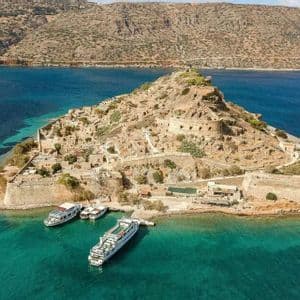 Vista aérea de una pequeña isla fortificada con ruinas de piedra, con varias embarcaciones atracadas en el mar turquesa circundante.