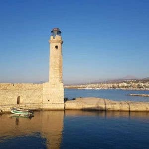Un faro de piedra se alza al final de un muelle junto a un mar azul en calma, con una ciudad portuaria visible al fondo.