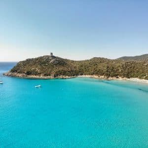 Vista aerea di una baia costiera con acqua turchese vivace, una spiaggia di sabbia e una torre di avvistamento in pietra su una collina verde.