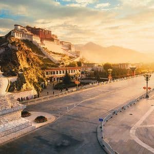 Un grande palazzo sorge su una collina rocciosa che domina un'ampia strada deserta all'alba, con uno stupa di pietra bianca in primo piano.