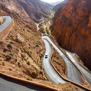 Vista aérea de un coche blanco en una carretera sinuosa con múltiples curvas cerradas, en un profundo cañón de roca roja.