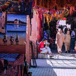 Un soleado callejón de mercado con gente paseando entre puestos de pinturas, cerámica y textiles coloridos.