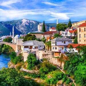 A coastal town with red-tiled roofs and a stone bell tower on the edge of a turquoise bay, with mountains in the distance.