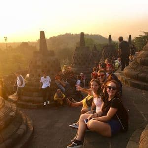Un voyage en groupe WeRoad posant pour une photo assis parmi des stupas de pierre lors d'un lever de soleil doré, avec un paysage forestier en arrière-plan.