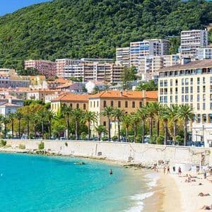 Una soleggiata cittadina costiera con palme ed edifici adagiati su una collina verde, che si affaccia su una spiaggia di sabbia e un mare turchese.