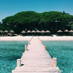 Un molo di legno si estende su acque limpide color turchese verso una spiaggia sabbiosa costeggiata da ombrelloni di paglia e alberi verdi lussureggianti.