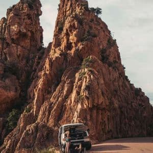 Un furgone bianco con il cofano aperto è parcheggiato su una strada di montagna ai piedi di una grande formazione rocciosa rossa, con due persone nelle vicinanze.