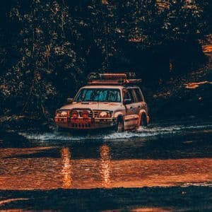 Un veicolo fuoristrada bianco attraversa un fiume poco profondo, schizzando acqua, con una fitta foresta sullo sfondo al tramonto.