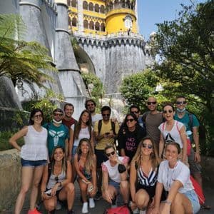 Un viaje en grupo de WeRoad sonríe para una foto grupal frente a un colorido castillo amarillo y gris bajo un cielo azul claro.