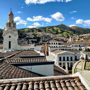 Una vista sobre los tejados de terracota de una ciudad con una torre de iglesia blanca y un edificio abovedado, con una montaña verde de fondo bajo un cielo azul.