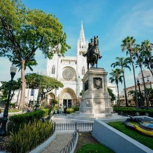 Una estatua ecuestre se alza en un parque de la ciudad frente a una gran catedral blanca, rodeada de palmeras y vegetación.