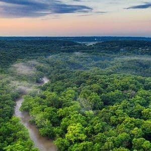 Una vista aérea de un río serpenteando a través de un vasto bosque verde con neblina baja bajo un cielo al atardecer.
