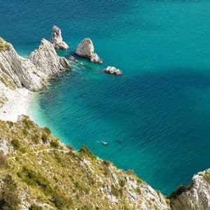 Veduta dall'alto di una costa rocciosa con ripide scogliere bianche, una spiaggia isolata e grandi formazioni rocciose in acqua turchese.