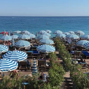 File di ombrelloni a strisce bianche e blu su una pedana di legno dove le persone si rilassano su lettini in riva al mare.