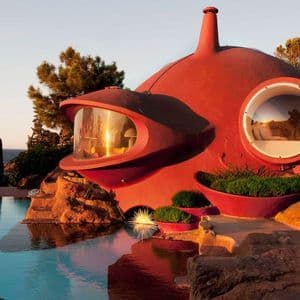 Una futuristica casa a bolla rossa con ampie finestre si erge su una scogliera rocciosa accanto a una piscina al tramonto.
