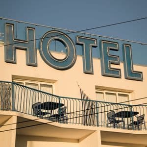 Una vista dal basso della facciata di un hotel con una grande insegna blu retrò sopra balconi con tavoli e sedie sotto un cielo sereno.